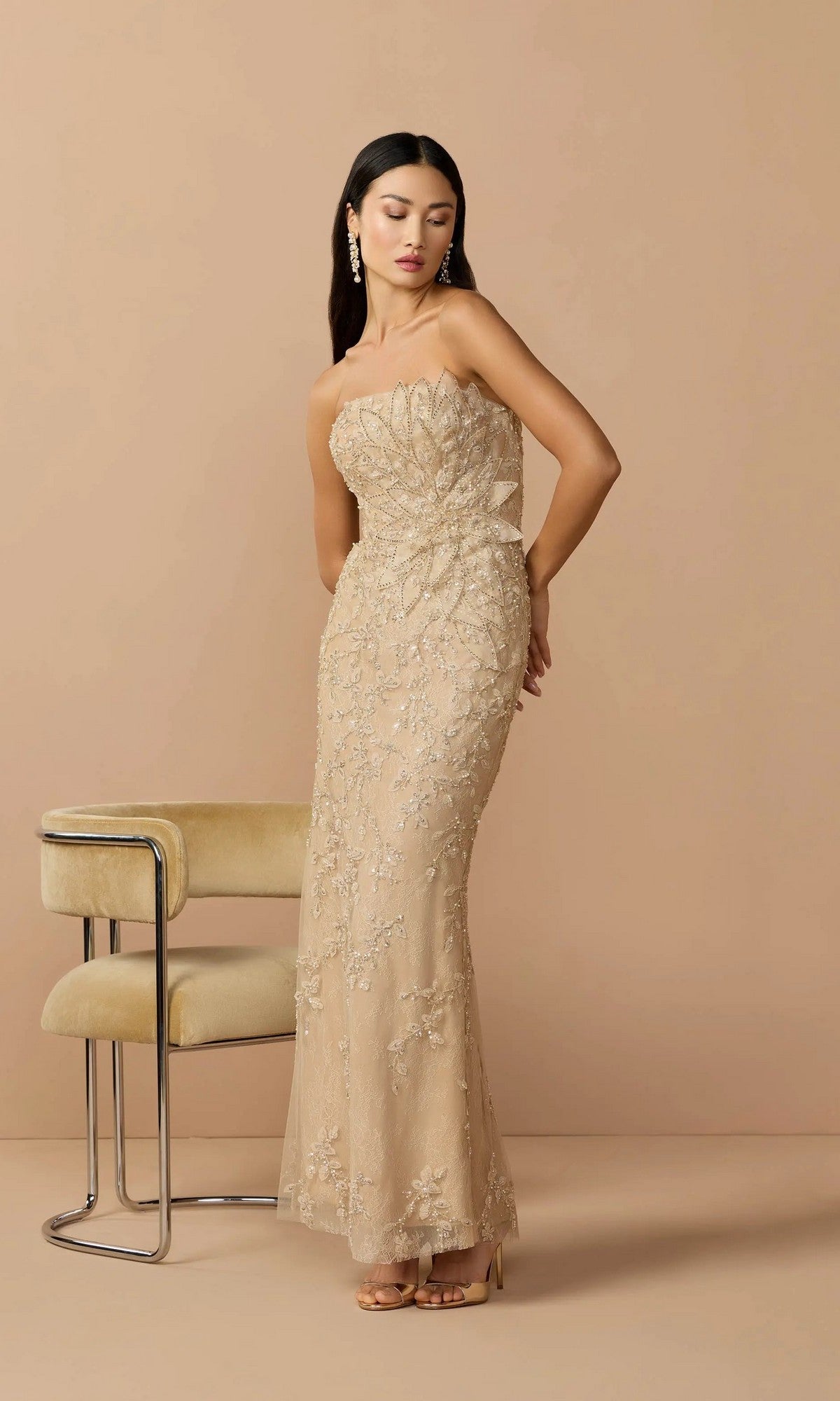 A woman wears the Mac Duggal 21200 long formal dress—a gold strapless gown with intricate beading—standing beside a light-colored chair against a plain beige background.